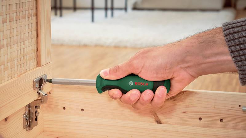 Eine Hand benutzt einen grünen Schraubendreher, um ein Scharnier an einem Möbelstück aus Holz festzuziehen. Der Hintergrund zeigt ein Interieur eines Raums mit hellem Ton.
