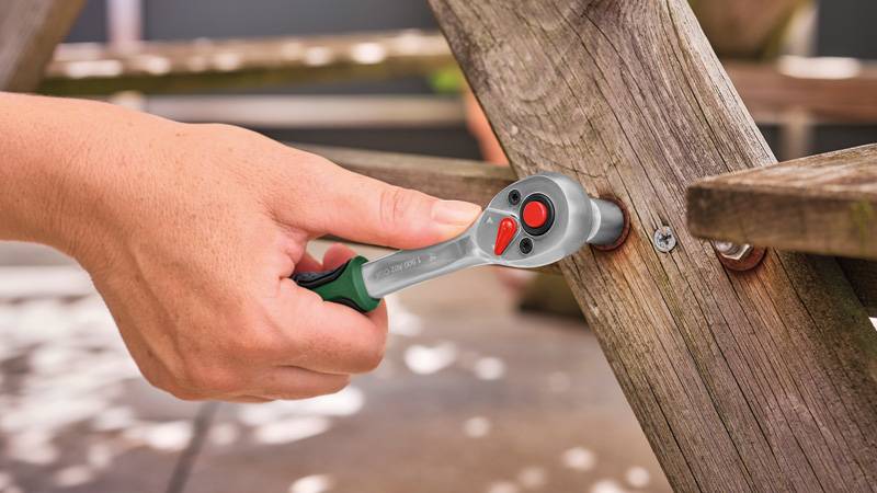 Eine Hand verwendet einen Ratschenschlüssel, um eine Schraube an einer Holzkonstruktion im Freien anzuziehen und demonstriert dabei die Werkzeugverwendung zum Zusammenbau oder zur Reparatur.