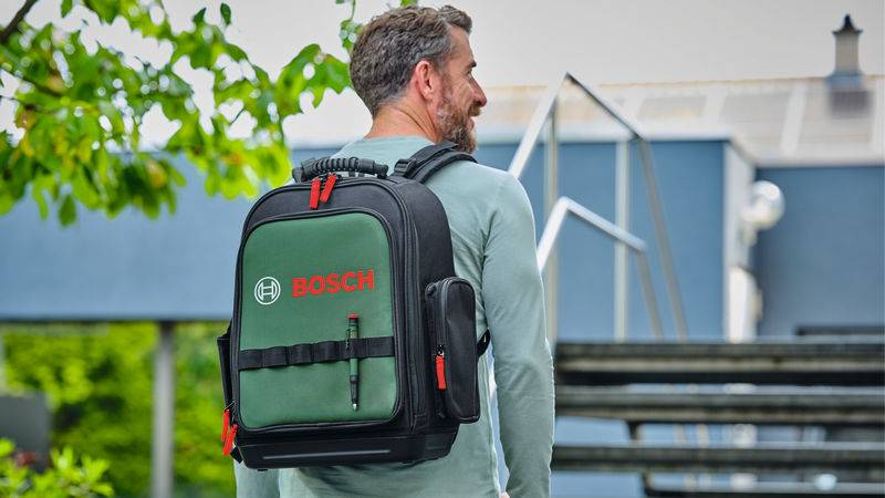 Ein bärtiger Mann trägt einen grün-schwarzen Rucksack mit Bosch-Markenlogo. Er ist im Freien, in der Nähe einer Treppe, mit Grünflächen im Hintergrund.