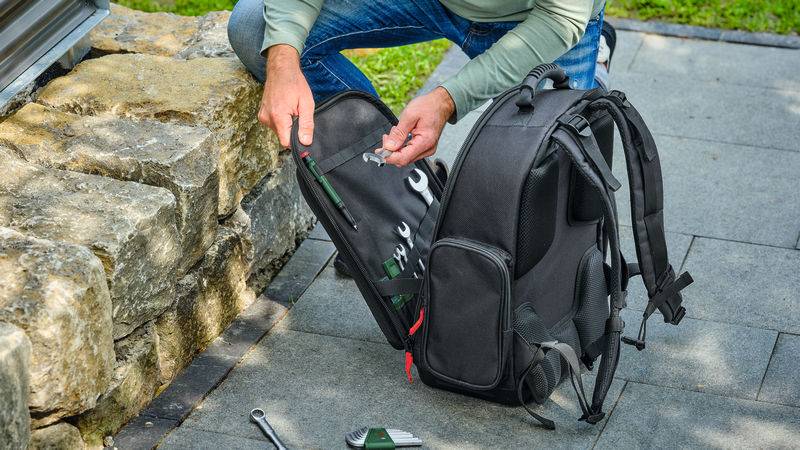 Eine Person kniet neben einer Steinmauer und ordnet Werkzeuge in einem schwarzen Rucksack auf einer gepflasterten Oberfläche an, mit Grünflächen im Hintergrund.