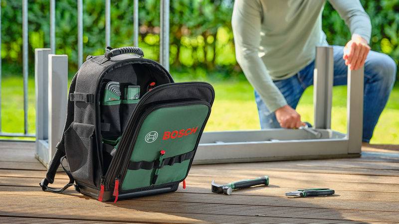 Eine Person montiert Möbel auf einer Holzterrasse mit Werkzeugen. Ein schwarzer und grüner Rucksack mit dem Logo von „Bosch