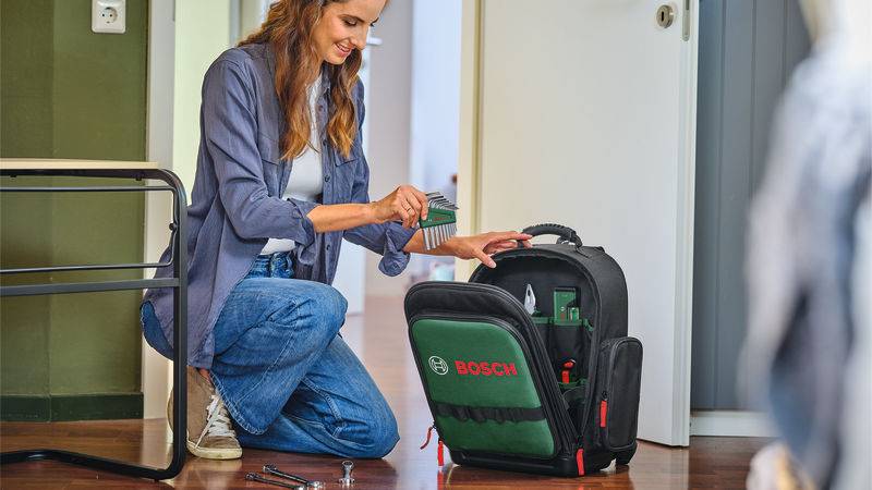 Eine Person kniet auf dem Boden und legt einen kabellosen Schrauber in eine grüne Bosch-Werkzeugtasche in einer häuslichen Umgebung.