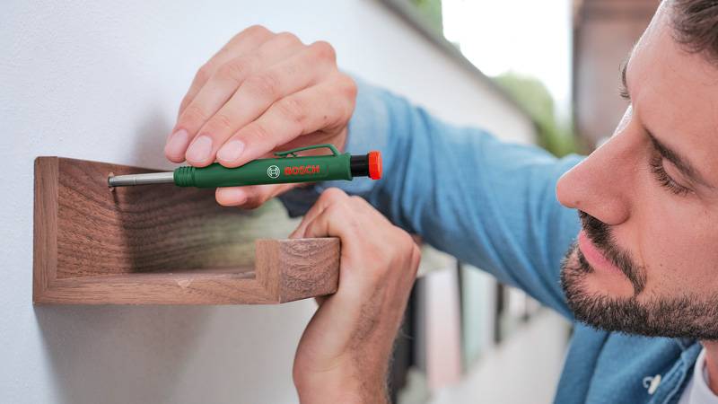 Eine Person verwendet einen grünen Schraubendreher, um ein hölzernes Regal an einer weißen Wand anzubringen. Der Fokus liegt auf der präzisen Handarbeit.