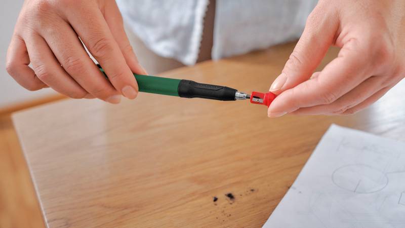 Zwei Hände montieren einen grünen Druckbleistift, indem sie eine rote Bleimiene einführen, während Zeichenpapier auf einem Holztisch in der Nähe liegt.