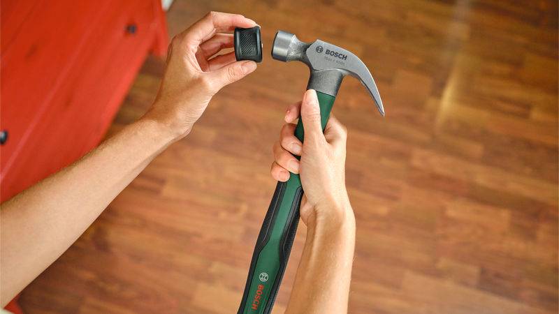 Eine Person hält einen Hammer und stellt ein kleines Gerät daran ein, in einem Raum mit Holzfußboden, was auf eine Werkzeug- oder technologiebezogene Aufgabe hindeutet.