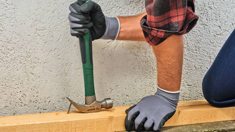 Eine Person mit Handschuhen sichert mit einem grünen Hammer eine Holzplanke gegen eine raue Wand.