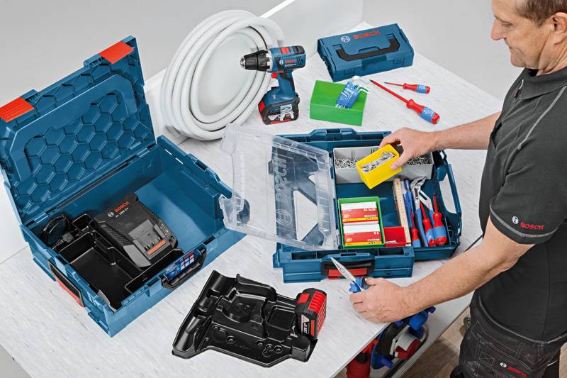 Eine Person organisiert Werkzeuge und Materialien wie einen Bohrer, ein Ladegerät, Kabel und Batterien in einer blauen Werkzeugbox auf einem weißen Tisch.