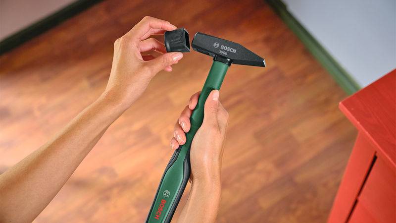 Eine Person verwendet ein Bosch-Multifunktionswerkzeug mit einer Schleifaufsatz über einem Hartholzboden, neben einem roten Möbelstück.