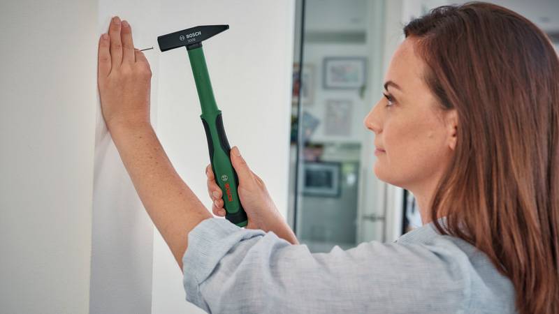 Eine Person verwendet einen Hammer mit grünem Griff, um einen kleinen Nagel in eine weiße Wand zu schlagen, wobei sie sich auf die Aufgabe in einem gut beleuchteten Raum konzentriert.