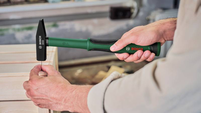 Eine Person verwendet einen grünen und schwarzen Bosch-Hammer, um einen Nagel in ein Holzbrett zu treiben und demonstriert damit Heimwerkerarbeiten oder Zimmermannswerk.