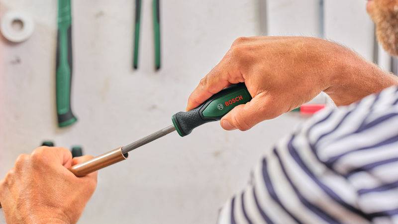 Eine Person in einem gestreiften Hemd benutzt einen Bosch-Schraubendreher, um an einem zylindrischen Metallobjekt zu arbeiten, während Werkzeuge im Hintergrund an der Wand hängen.