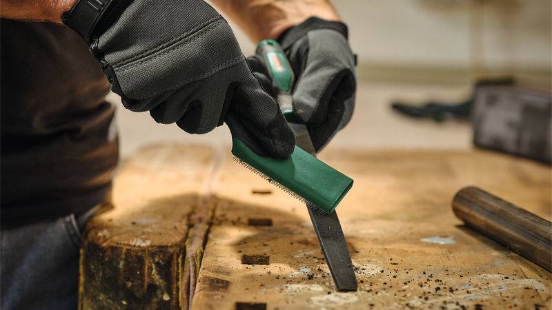 Eine Person mit schwarzen Handschuhen schärft ein Werkzeug mit grünem Griff auf einer Werkbank und konzentriert sich auf die Kante des Werkzeugs zum Feinabstimmen.