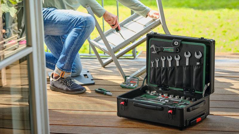Eine Person montiert Gartenmöbel auf einer Holzterrasse mit Werkzeugen aus einer offenen Werkzeugkiste, mit grünem Rasen im Hintergrund sichtbar.