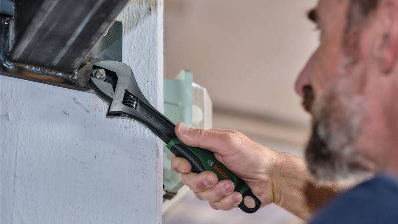 Ein Mann verwendet einen Schraubenschlüssel, um eine Schraube an einem Metallträger zu befestigen, der an einer Wand angebracht ist, und demonstriert dabei Wartungs- oder Reparaturarbeiten.