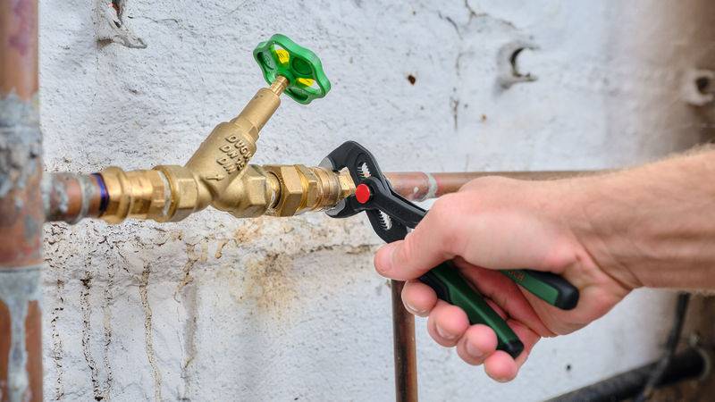 Eine Person verwendet eine Zange, um ein Messingventil an einem Kupferrohr gegen eine weiße Betonwand festzuziehen, was auf Klempnerarbeiten hindeutet.