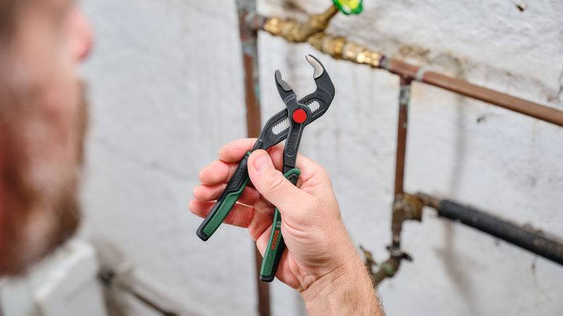 Eine Person, die mit einer Zange eine Rohrverbindung gegen eine weiße Wand festzieht. Werkzeuge und Klempnerarbeiten sind im Vordergrund sichtbar.