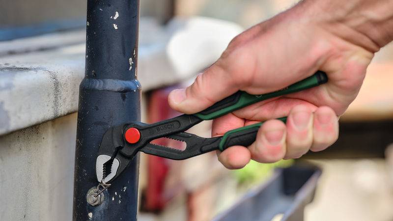 „Eine Hand benutzt eine Zange, um ein rundes Metallobjekt auf einem Rohr zu greifen und zu drehen, wahrscheinlich bei einer Wartungs- oder Reparaturarbeit im Freien.