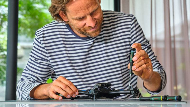 Eine Person in einem gestreiften Hemd repariert eine Drohne auf einem Tisch mit einem Schraubenzieher, während Bäume außerhalb durch das Fenster sichtbar sind.