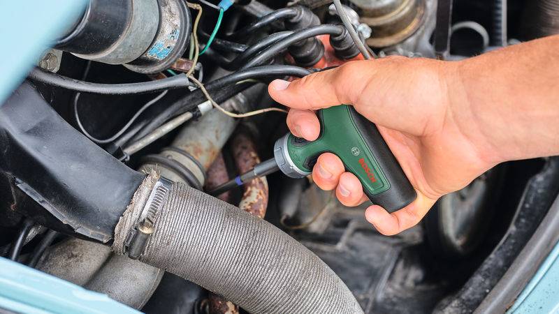 Eine Hand hält ein grünes Werkzeug mit Bosch-Logo und justiert oder arbeitet an Motorteilen im Motorraum eines Autos.