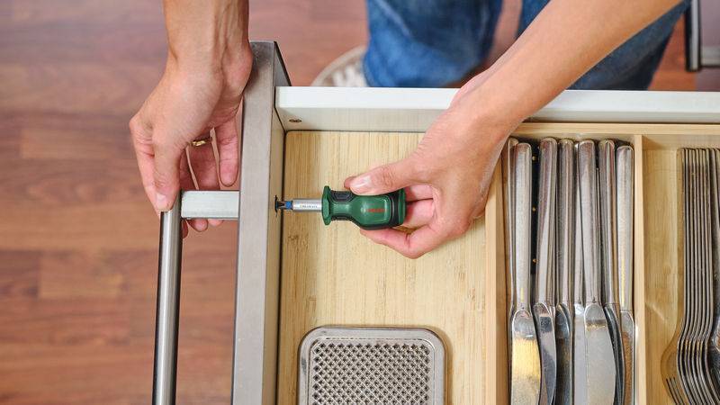 Eine Person zieht einen Küchenschubladengriff mit einem Schraubenzieher fest; die Schublade enthält Utensilien und Besteck auf einem hölzernen Organizer.