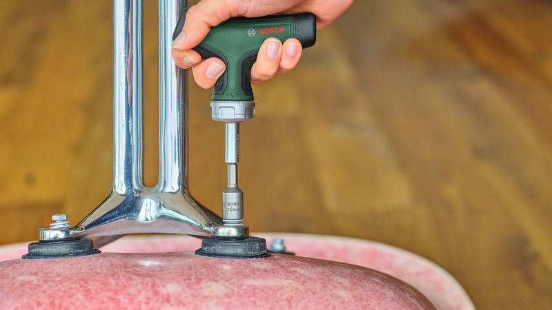 Eine Hand benutzt ein Bosch-Elektrowerkzeug, um eine Schraube an einem Metallstuhlbein festzuziehen, das an einem rosa Stuhl befestigt ist.
