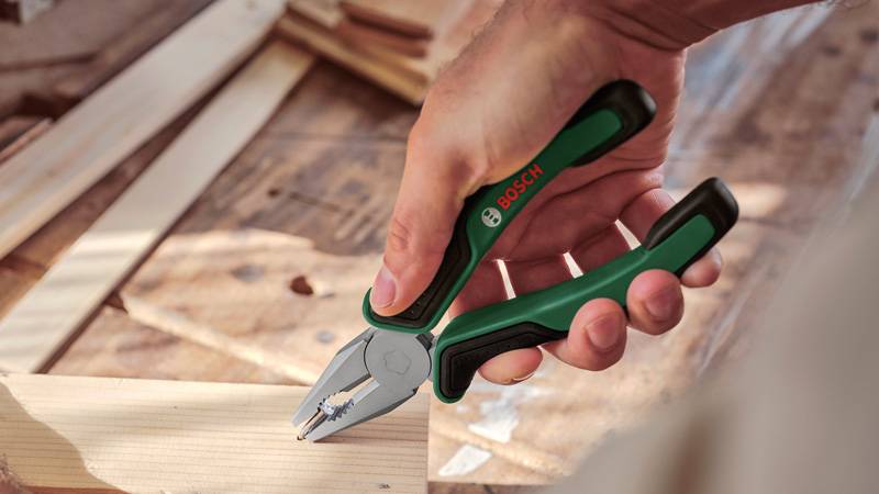 Eine Hand hält ein Paar grün-schwarzer Zangen über einer Holzoberfläche, wobei natürliches Licht die Szene beleuchtet.