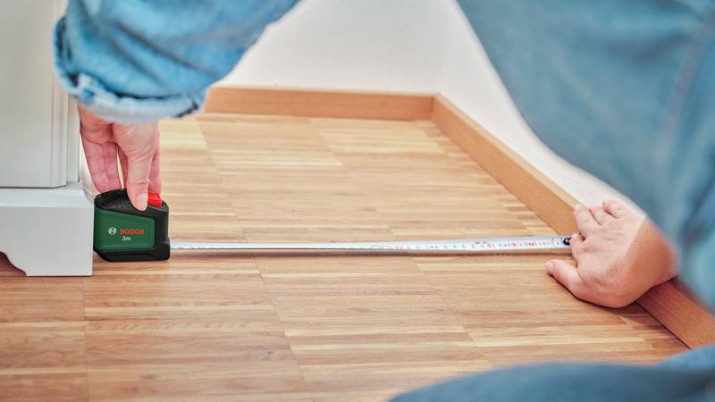 Eine Person nutzt ein Stahllineal, um die Entfernung zwischen einer Wand und einem Gerät auf einem Holzboden zu bestimmen.
