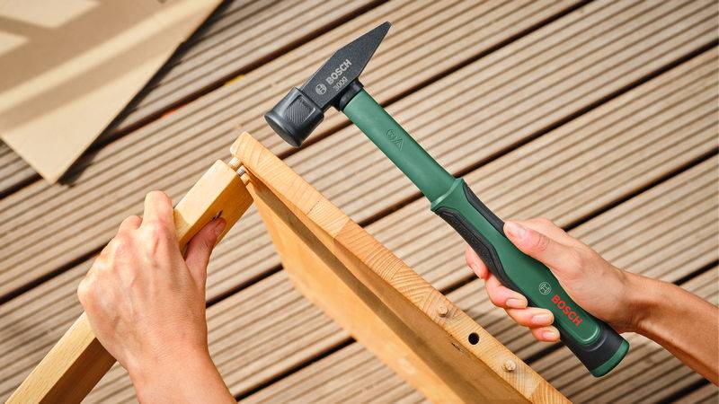 Eine Person montiert Möbel aus Holz auf einer Holzterrasse mit einem Hammer.