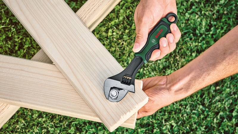 Eine Person verwendet einen verstellbaren Schraubenschlüssel, um eine Schraube an Holzbalken zu festigen, die auf Gras platziert sind.