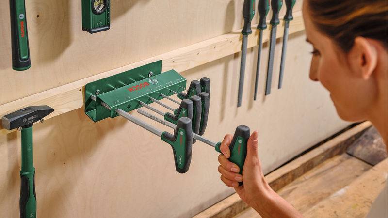 Eine Person organisiert und lagert Schraubendreher in einem grünen wandmontierten Gestell in einer Werkstatt. Weitere Werkzeuge sind ebenfalls an der Wand sichtbar.