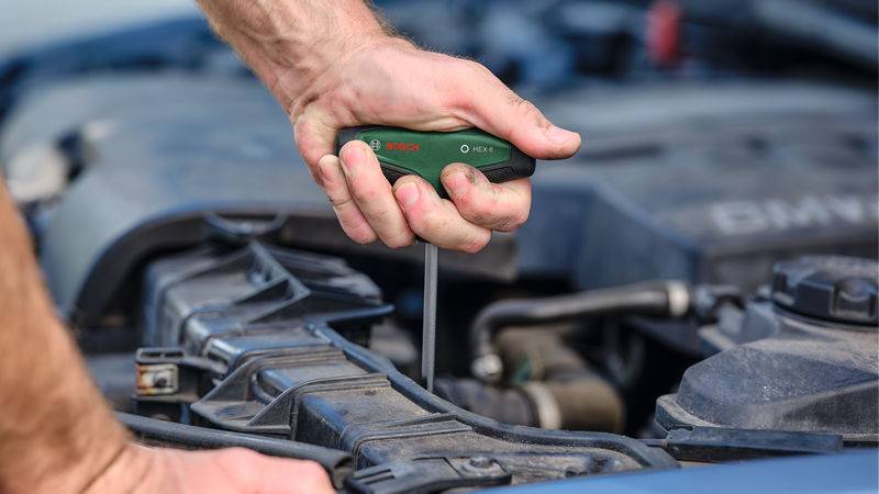 Eine Person verwendet einen grünen Bosch-Schraubenzieher, um das Motorkompartiment eines Fahrzeugs einzustellen oder zu reparieren und konzentriert sich dabei auf detaillierte Handarbeit.