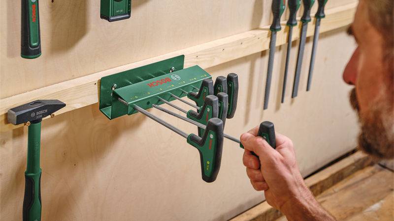 Eine Person ordnet lange Schraubendreherbits an einem an der Wand montierten Werkzeughalter neben einem Hammer und anderen Werkzeugen in einer Werkstattatmosphäre an.