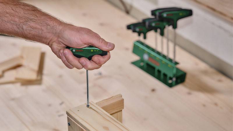 Eine Person verwendet einen grünen Schraubendreher, um eine Schraube in ein Holzstück zu treiben, wobei weitere Werkzeuge und ein Holzboden im Hintergrund sichtbar sind.