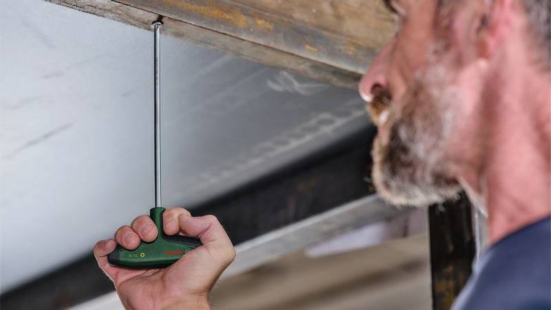 Eine Person, die einen langen Schraubendreher verwendet, um etwas auf einer Deckenoberfläche zu reparieren oder anzupassen, wobei der Fokus auf dem Werkzeug und der anstehenden Aufgabe liegt.
