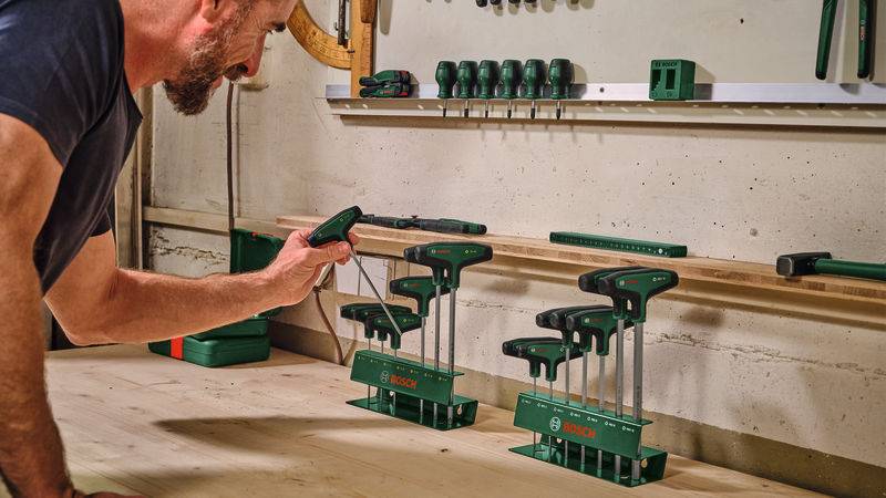 Eine Person organisiert Bosch-Handwerkzeuge in einer Werkstatt und ordnet sie ordentlich in Haltern auf einer Holzbank an, während andere Werkzeuge im Hintergrund zu sehen sind.