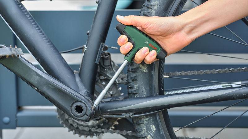 Eine Person verwendet einen T-Griff-Schlüssel, um die Kurbel-Schraube an einem Fahrrad einzustellen. Das Fahrrad ist dunkelfarben, und das Werkzeug hat einen grünen Griff.