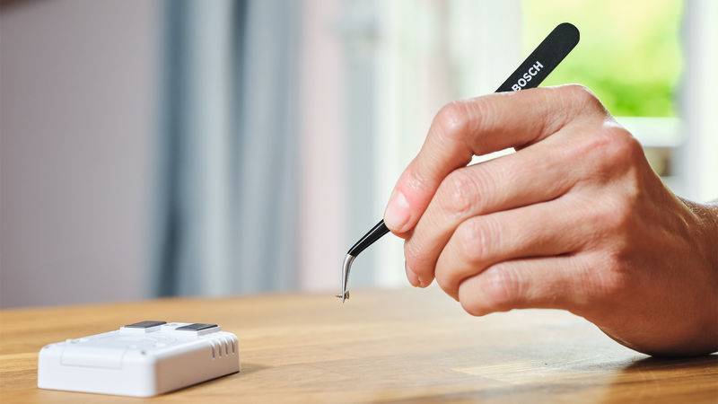 Eine Hand, die eine Pinzette hält, platziert eine kleine elektronische Komponente auf einem Holztisch neben einem weißen Gerät, wahrscheinlich im Zusammenhang mit Montage oder Reparatur.