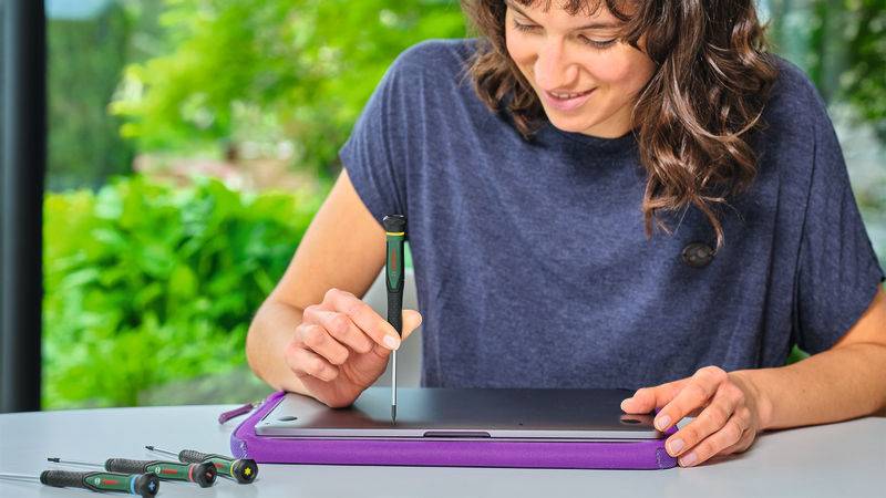 Eine Person lächelt, während sie ein Tablet mit einem Schraubenzieher an einem Tisch repariert, mit anderen Werkzeugen in der Nähe sichtbar, Grünpflanzen im Hintergrund.