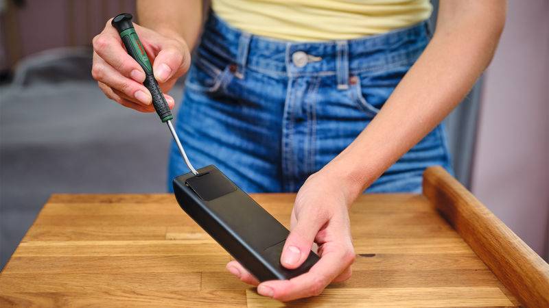 Eine Person benutzt einen Schraubendreher, um ein schwarzes Batteriefach auf einem Holztisch zu öffnen. Der Fokus liegt auf den Händen und dem Gerät.