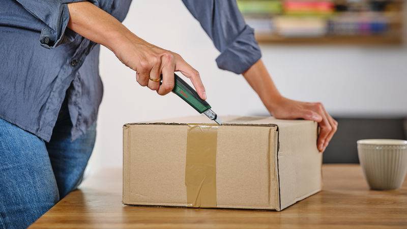 Eine Person schneidet eine versiegelte Pappschachtel auf einem Holztisch auf, neben einer Tasse. Regale mit bunten Büchern im Hintergrund.