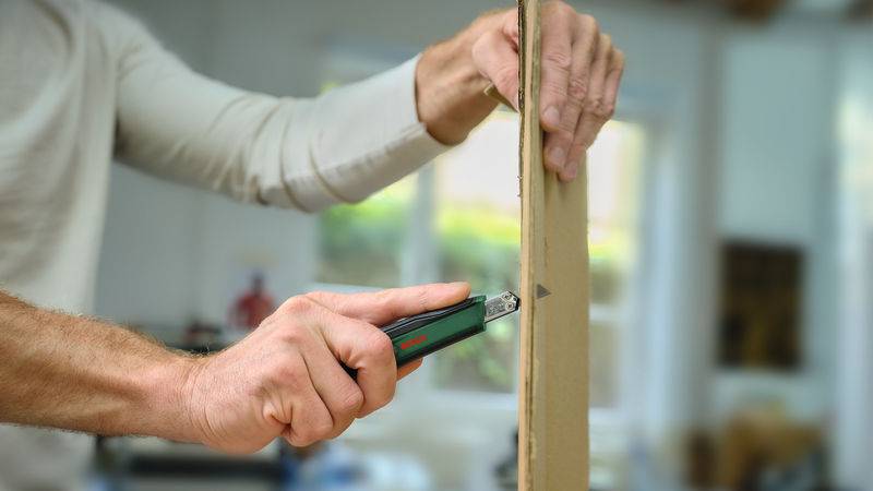 Eine Person benutzt ein Universalmesser, um ein Stück Pappe in einem hellen Raum sorgfältig zu schneiden und konzentriert sich dabei auf Präzision und Sicherheit.