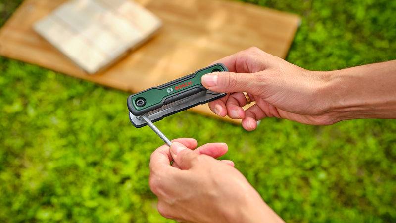 Eine Person verwendet einen Multitool-Schraubendreher an einem sonnigen Tag, mit einem Holzbrett und Gras im Hintergrund.