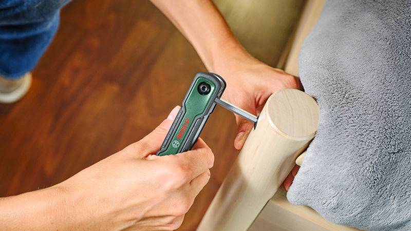 Person, die einen grünen Bosch-Elektroschrauber verwendet, um eine Schraube in einem Möbelstück aus Holz neben einem grauen Stoff festzuziehen.