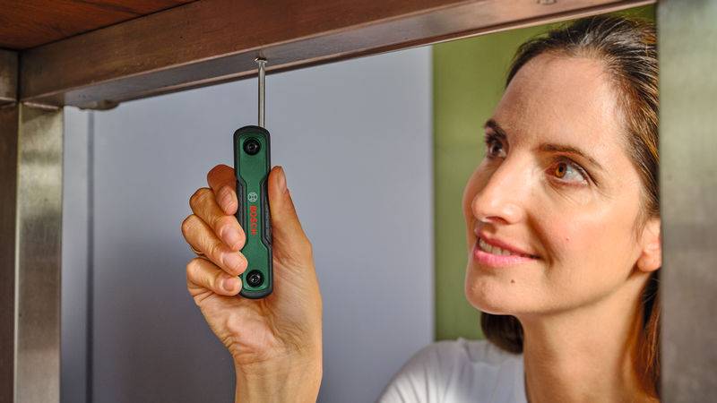 Eine Person verwendet ein grünes Bosch-Werkzeug, um eine Schraube unter einer Holzoberfläche festzuziehen, wobei sie sich intensiv auf die Aufgabe konzentriert.