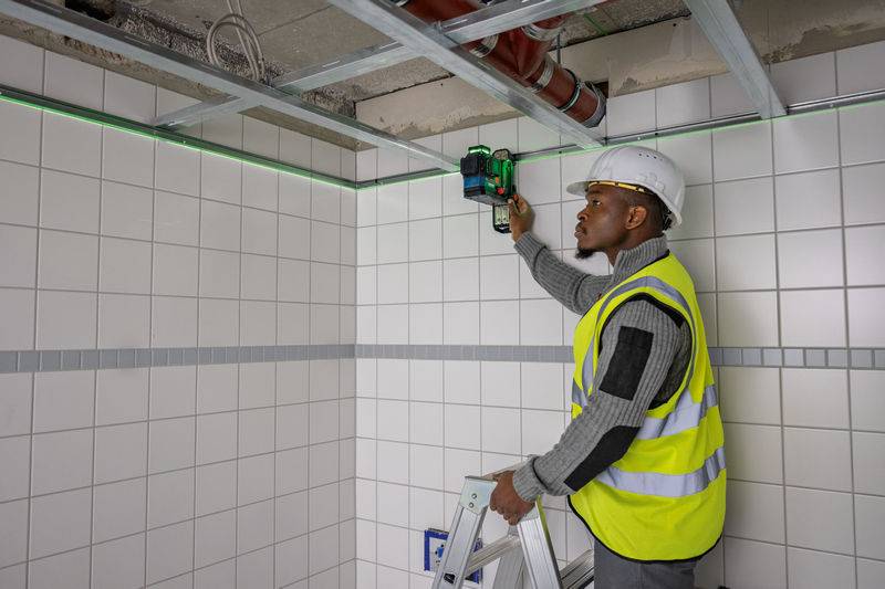 Ein Bauarbeiter, der eine Sicherheitsweste und einen Helm trägt, verwendet ein Laserniveau an einer gefliesten Wand und steht auf einer Stehleiter in einem unfertigen Raum.