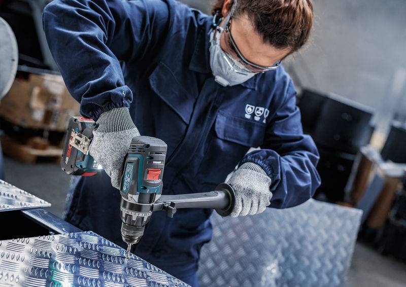 Eine Person in Schutzausrüstung bedient einen Elektroschrauber auf einem Metallblech in einer Industrieumgebung.