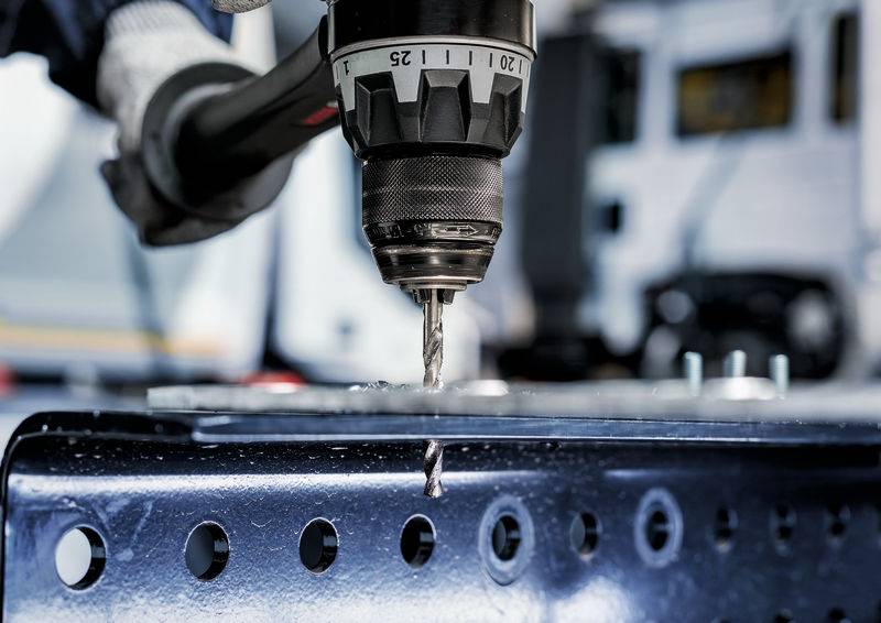 Eine Nahaufnahme einer Bohrmaschine, die ein Loch in ein Metallblech bohrt, wobei eine behandschuhte Hand sie stabilisiert. Ein unscharfer Industriehintergrund ist sichtbar.