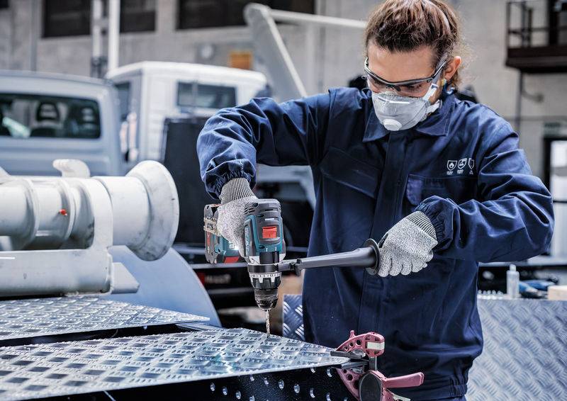Eine Person in Schutzausrüstung bedient eine Elektrobohrmaschine auf einem Metallblech in einer industriellen Umgebung und konzentriert sich dabei auf Präzision und Sicherheit.