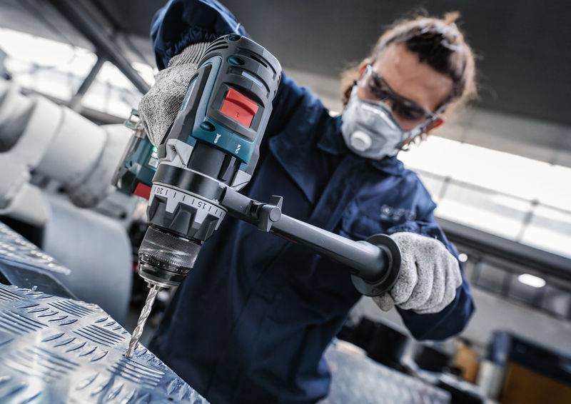 Eine Person, die Schutzausrüstung trägt, bedient eine Elektrobohrmaschine auf einer Metalloberfläche in einer industriellen Umgebung.