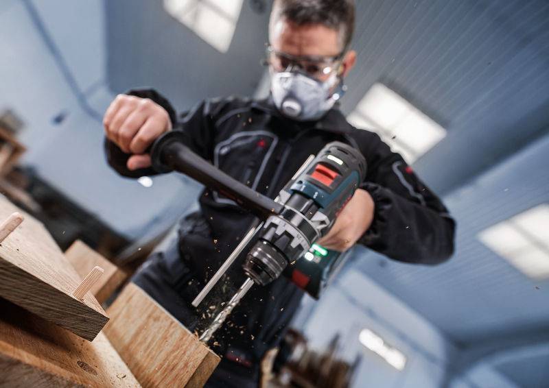 Eine Person, die eine Schutzbrille und eine Maske trägt, bedient einen Elektroschrauber an einem Holzstück, wobei Holzstaub in einer Werkstatt fliegt.